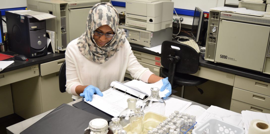Woman in hijab and rubber gloves looks through paperwork in lab