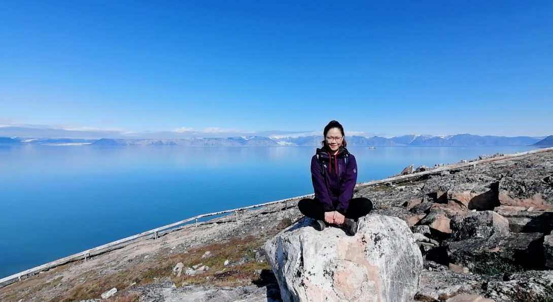 Cynthia sitting on a rock along the artic circle.  There is lake water and mountains in the background.