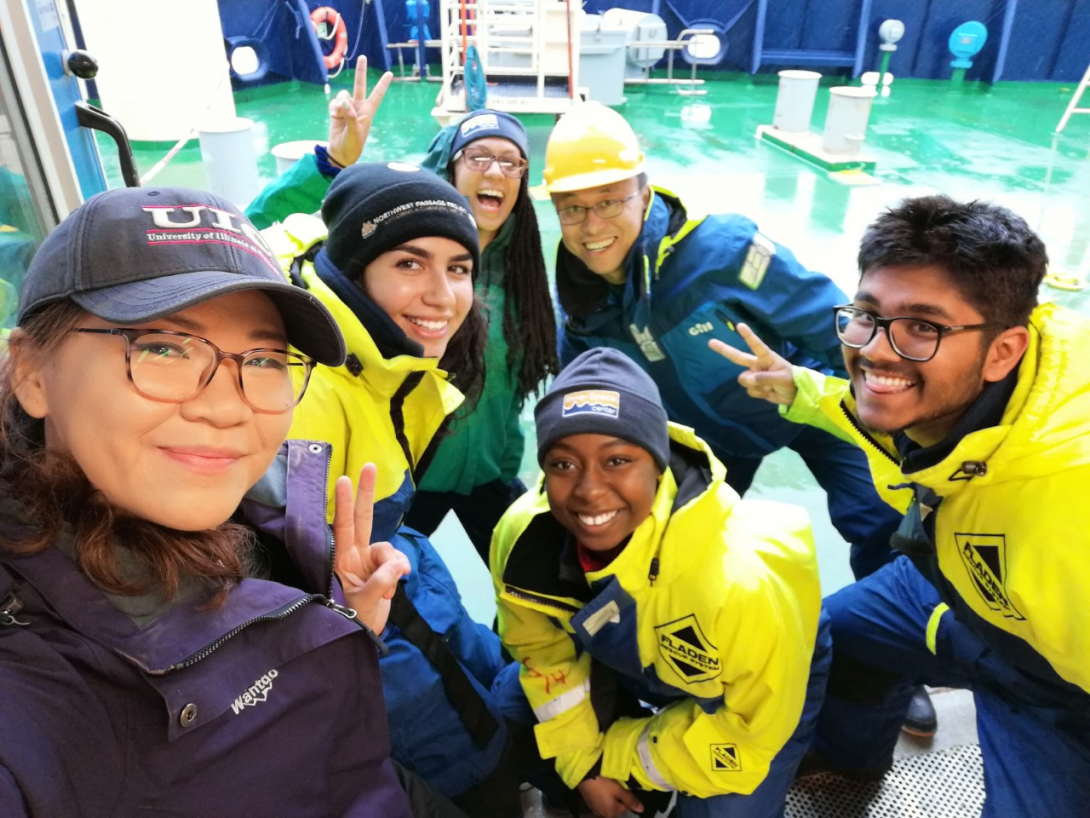 Group of 6 students  in front of lab equipment. Students are dressed with hats and big jackets.