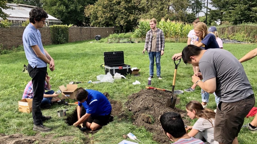 Group of geoscience students scatterd in a green gass field taking a soil sample. Some students have shovels, a hole is clearly dug and one student is sitting in it.