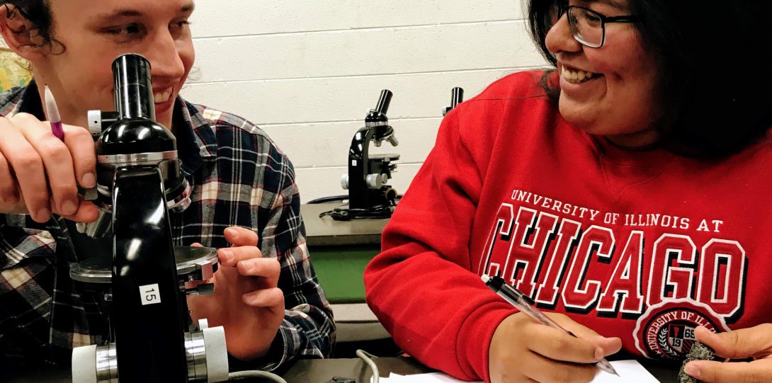 Two students collaborating in a classroom with a microscope and rock samples.