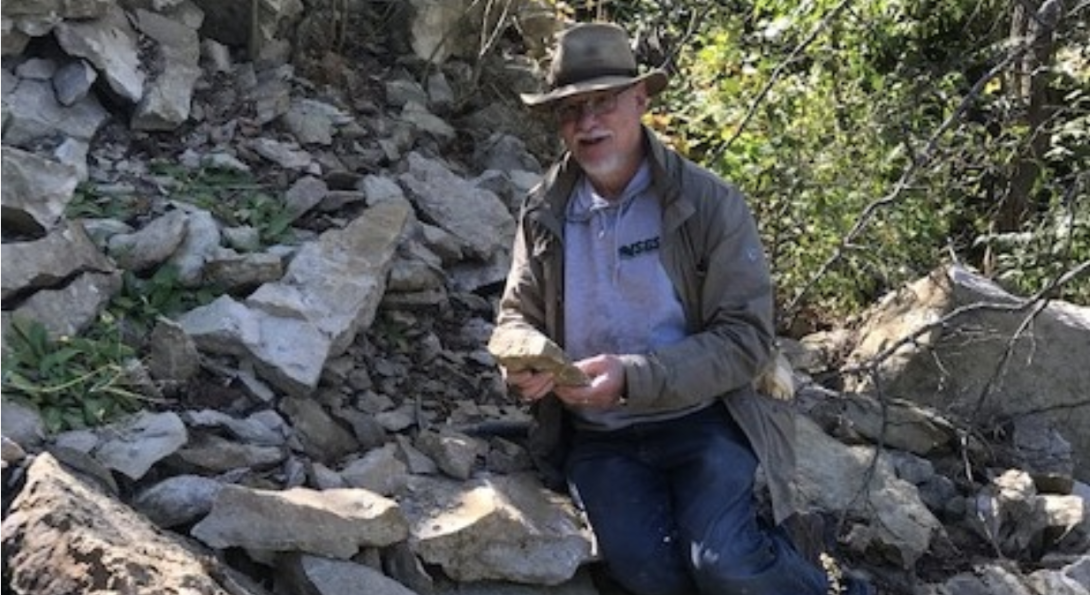 Man with hat on and rock sample in hand standing in front of loose rocks on an outcrop.