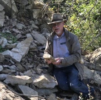 Man with hat on and rock sample in hand standing in front of loose rocks on an outcrop.
                  