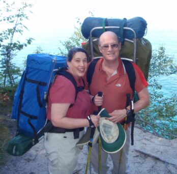 Spouses, husband and wife wearing large camping backpacks in an outdoor setting with vegetation. 