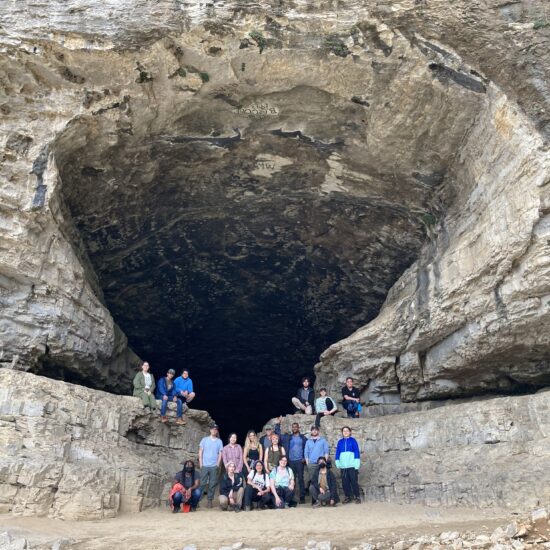 Group of students and faculty wearing different colored cloths standing in front of a large rock outcrop that features a large entrance to a cave